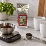 Ground coffee measured in a bowl on a kitchen scale beside an ANASU bag and two mugs